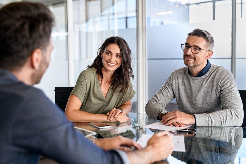 Zwei Geschäftspartner im Gespräch mit einem Berater in einem modernen Büro. Die beiden hören aufmerksam zu und lächeln.